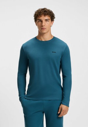 Långärmad skjorta i teal med rund halsringning, texturerat material och en liten svart logotyp på bröstet. Matchas med byxor i samma färg.