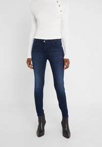 Jeans oscuros ajustados de mezclilla con una textura suave, cintura media, y bolsillos frontales, combinados con un suéter blanco de canalé ajustado y botas negras hasta el tobillo.