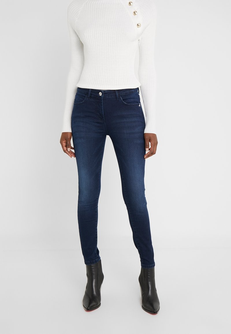 Jeans oscuros ajustados de mezclilla con una textura suave, cintura media, y bolsillos frontales, combinados con un suéter blanco de canalé ajustado y botas negras hasta el tobillo.