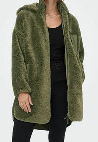 Zelená fleecová bunda s kapucí, s měkkým, strukturovaným povrchem. Má přední zip a boční kapsy, navlečená přes černý top.