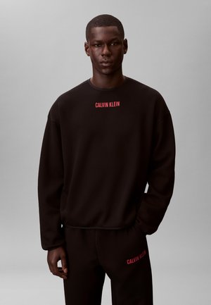 Młody mężczyzna ubrany w czarną bluzę i spodnie Calvin Klein z czerwonym napisem logo, stojący na jednolitym szarym tle.