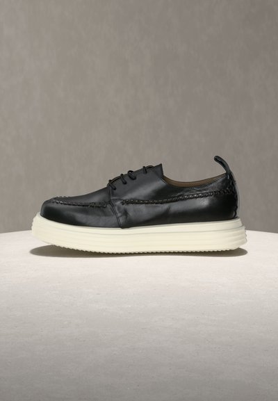 Μαύρο δερμάτινο παπούτσι με κορδόνια, χοντρή λευκή σόλα, ραφές και ταινία τράβηγματος σε ουδέτερο γκρι και μπεζ φόντο.