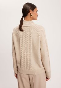 Beżowy sweter dzianinowy z wysokim kołnierzem, prążkowanymi rękawami i teksturowanym wzorem w warkocz na plecach, zestawiony z jasnobeżowymi spodniami.