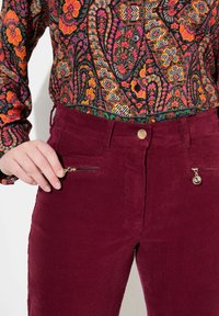 Pantaloni in velluto a coste bordeaux con tasche a zip dorate e chiusura con bottone, abbinati a una camicia a fantasia floreale colorata con accenti neri, rossi e verdi.