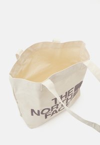 Bolso de lona en beige claro con dos asas largas; presenta un gran logo impreso en marrón oscuro; parte superior abierta con interior sin forrar.