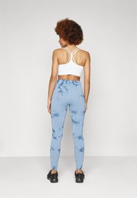 Brassiere sportivo bianco con design incrociato sulla schiena, abbinato a leggings azzurri tie-dye con texture morbida e scarpe da ginnastica nere.
