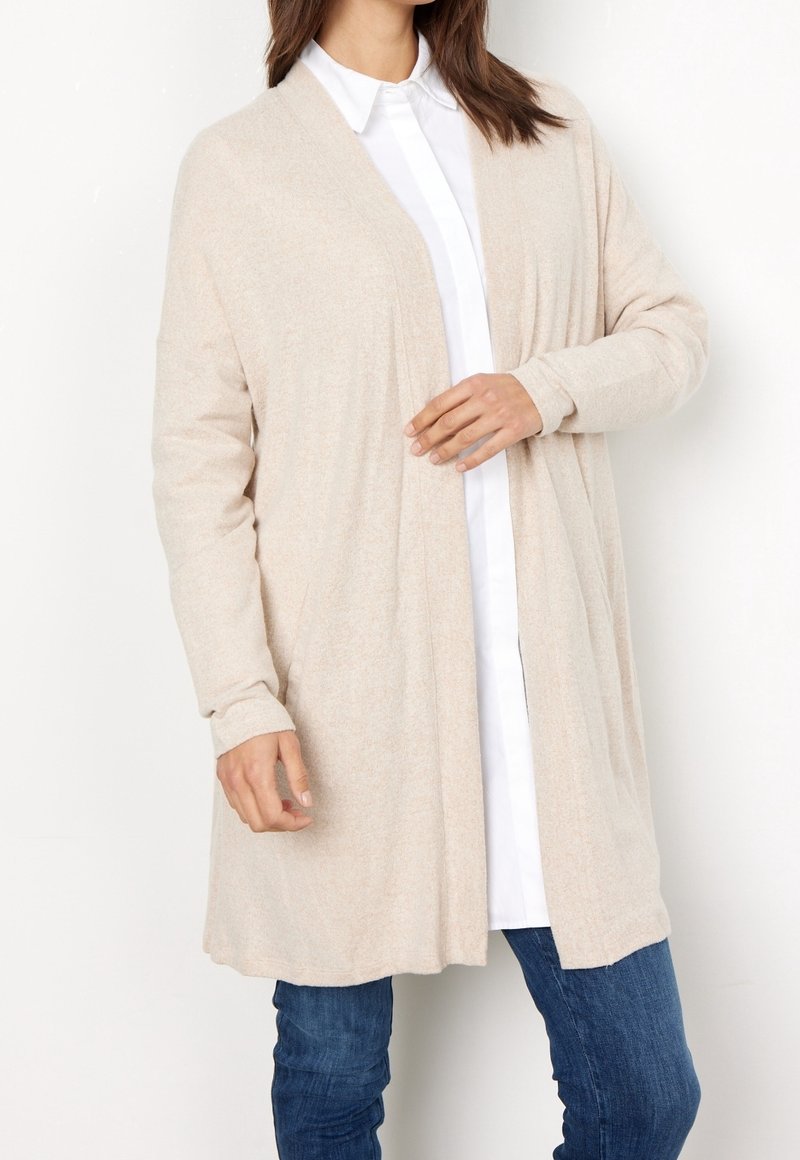 Gilet beige clair avec une ouverture sur le devant, de longues manches et une texture douce, porté sur une chemise blanche à boutons. Design décontracté, sans fermer.