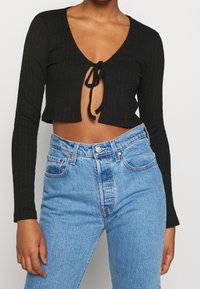 Svart ribbad crop top med långa ärmar och djup V-ringning, med en framknutning som detalj. Kombinerad med högmidjade blå denimjeans.
