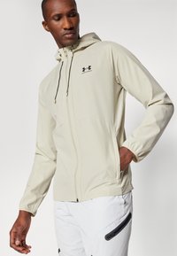 Giacca beige con cappuccio dotata di zip frontale, polsini elasticizzati e tasche laterali; presenta un logo nero sul petto; texture liscia.