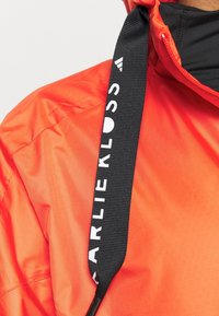 Personne portant une veste orange vif avec cordon de capuche noir marqué "KARLIE KLOSS" en texte blanc, gros plan sur l'épaule et le col.