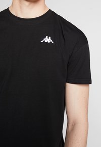 Černé bavlněné tričko s krátkým rukávem, s vyšitým bílým logem na levé hrudi. Hladká textura, kulatý výstřih.