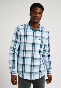 Koszula z długim rękawem, zapinana na guziki, w niebiesko-czarny wzór w kratkę, z bawełnianego materiału, z lekko zaokrąglonym dołem oraz małym logo na kieszeni piersiowej.