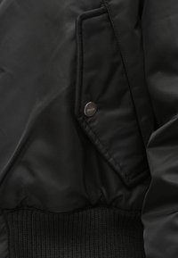 Svart nylon bomberjakke med ribbet kant, klaffelomme med knapp og glatt tekstur. Minimalistisk design med subtil merking.