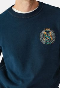 Felpa in cotone blu navy con collo rotondo, caratterizzata da logo ricamato in oro, verde e bianco, e una finitura testurizzata.