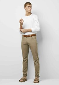 Weißes Langarmhemd, khakifarbene Hose mit Gürtel und gemusterte Sandalen. Das Modell steht aufrecht vor einem einfarbigen Hintergrund.