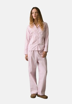 Mujer con un conjunto de pijama de botones verticales en rosa y blanco y zapatillas marrones, de pie contra un fondo liso claro.