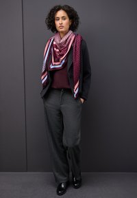 Blazer nero sopra un maglione bordeaux, pantaloni larghi grigi e una sciarpa fantasia nei colori rosa, bordeaux e blu. Stivali neri alla caviglia completano il look.