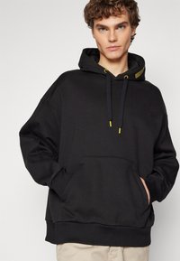 Felpa nera con un fit rilassato, lacci con accenti gialli, tasca frontale, polsini e orlo a coste, realizzata in materiale morbido.