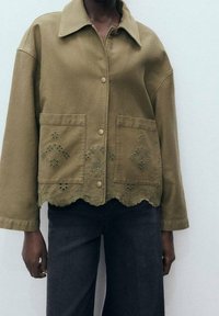 Olijfgroene denimjack met een kraag, gegolfde zoom en geborduurde diamantpatronen op de zakken. Voorzien van knoopsluitingen.