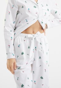 Biały komplet piżamy z wzorem nart i sosen. Koszula z długim rękawem zapinana na guziki, z kieszenią oraz spodnie na gumce związywane w pasie.