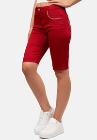 Bermuda shorts in denim rosso con un design fitted, dotati di una chiusura zip e due tasche frontali, una delle quali con una cerniera decorativa. Indossati con sneakers bianche.