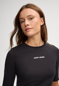 Haut ajusté à manches longues noir avec un col rond et le texte blanc "JOOP! JEANS" imprimé sur le devant. Texture de tissu lisse.