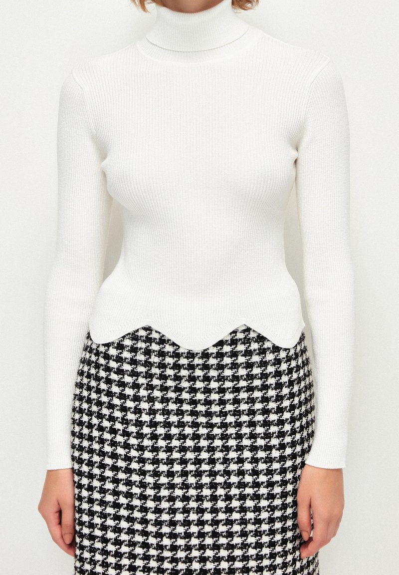 Pull à col roulé blanc côtelé avec un ourlet festonné, associé à une jupe crayon à motif pied-de-poule noir et blanc.