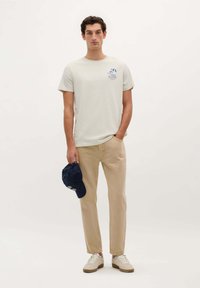 Helles graues T-Shirt für Herren mit einem kleinen blauen Grafikdruck, beigen geraden Hosen und einer marineblauen Kappe, ergänzt durch weiße Sneaker.