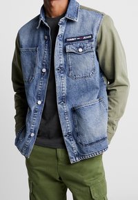 Farkkutakki, jossa on oliivinvihreät hihat, napit ja taskut; rinnassa on "TOMMY JEANS" -patch. Päällä musta paita.