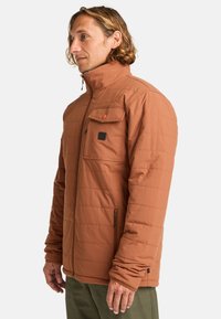Chaqueta aislante marrón con un alto cuello, textura acolchada, bolsillo en el pecho delantero y cremalleras laterales, con un parche de logo en el pecho.