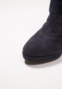 Gros plan sur la pointe d'une bottine en suède noir avec des surpiqûres visibles et une semelle plateforme basse sur un fond blanc.