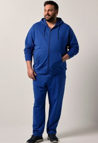 Uomo che indossa una felpa con zip di colore blu reale e pantaloni della tuta abbinati, in piedi con una mano in tasca e guardando di lato.