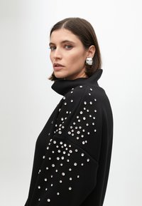 Schwarzer Pullover mit hohem Kragen und verstreuten silbernen Strasssteinen an den Ärmeln. Glatte Stofftextur, taillierte Passform, minimalistischer Stil.