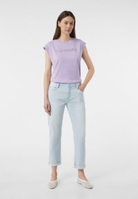 Helles Lavendel-T-Shirt mit dem Aufdruck "LAVENDER", kombiniert mit hellblauen Jeans mit hochgerollten Säumen und weißen Mesh-Slippern. Schlichte Gestaltung.