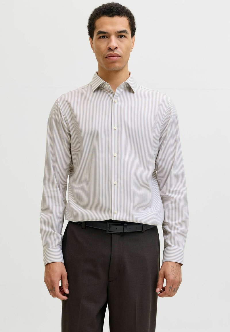 Chemise boutonnée avec des rayures verticales en beige clair et gris. Manches longues, col pointu, et coupe ajustée. Associée à un pantalon foncé.