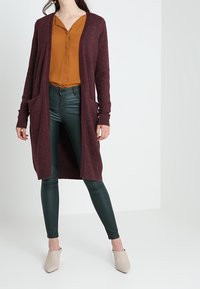 Burgundin pitkä neuletakki, jossa on taskut, puettua vaaleankeltaisen paidan ja vihreiden keinonahkahousujen päällä. Vaaleat teräväkärkiset korkokengät täydentävät asua.