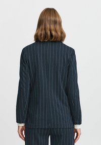 Tumesinine pinstripe bleiser, kohandatud disain, struktureeritud õlad, üks lõhe seljal ja valged pahkluud, mis piiluvad varrukatest välja.