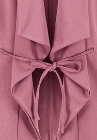 Tissu mauve avec col drapé attaché au centre par une ceinture nœud en tissu assorti, montrant des coutures détaillées.