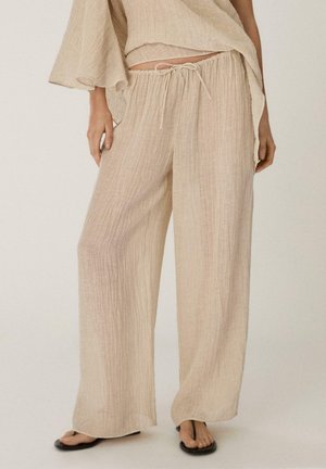 Kvinde iført løstsiddende, beige teksturerede snorebukser kombineret med matchende flydende top og sorte flip-flops, stående mod en ensartet baggrund.