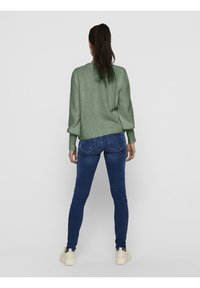 Grön texturerad tröja med långa ärmar, kombinerad med blå smala jeans och ljusa sneakers, sedd bakifrån.