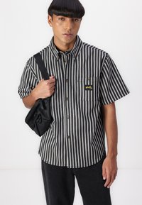 Short-sleeve, button-up shirt with black and white vertical stripes, featuring a left chest pocket and gold logo. Model holding a black bag.