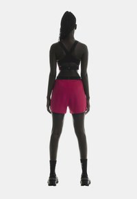 Sort sports-bh med udsnit i ryggen, parret med lyserøde shorts. Modellen bærer sorte sokker og teksturerede sorte sneakers. Enkel atletisk design.