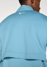 Jacheta Nike de culoare albastru deschis are un guler înalt, o textură netedă și un logo alb pe spate, evidențiind un design modern, stratificat.