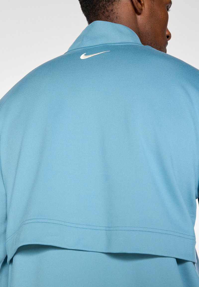 Giacca Nike azzurro chiaro con colletto alto, texture liscia e logo bianco sul retro, che mette in mostra un design moderno e stratificato.