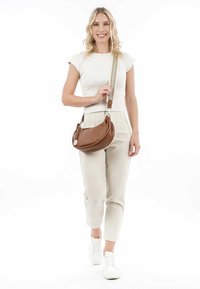 Brown leather crossbody bag with a rounded shape, textured finish, silver hardware, and adjustable strap, carried by a person in light clothing.