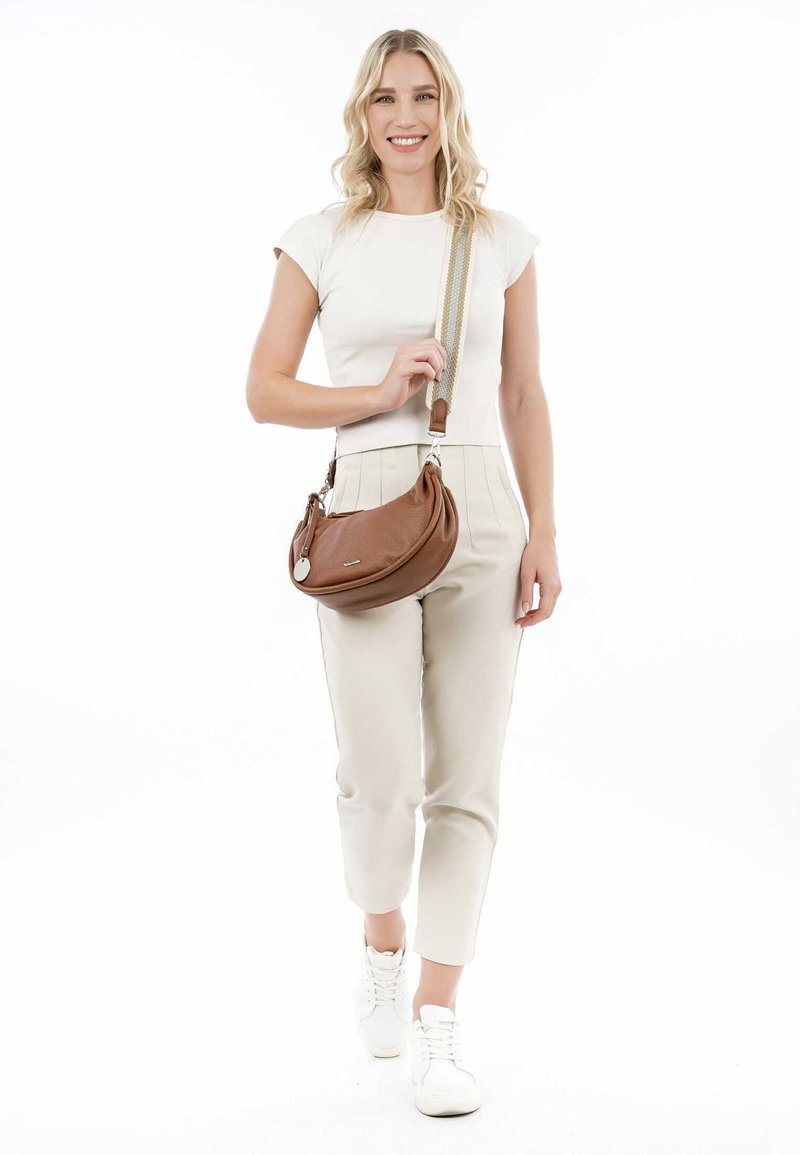 Brown leather crossbody bag with a rounded shape, textured finish, silver hardware, and adjustable strap, carried by a person in light clothing.