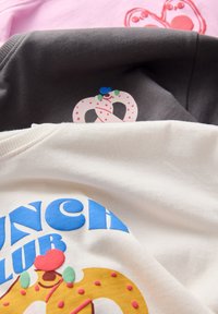 Trzy koszulki w tkaninach: białej, szarej i różowej. Każda z nich ma kolorowe grafiki: precel na szarej, napis "BRUNCH CLUB" na białej oraz wzór na różowej.