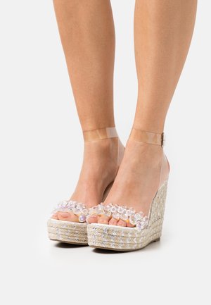 Sandales compensées avec une base en jute tissé, des brides en PVC transparent, ornées de grandes pierres précieuses transparentes à l'avant.