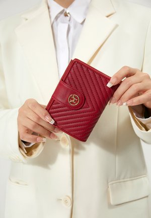 Manos con manicura francesa sosteniendo una cartera de cuero rojo acolchado con un cierre de logo dorado, vistiendo un blazer crema y camisa blanca.