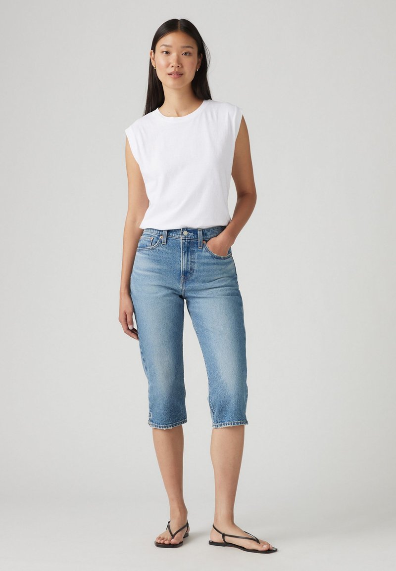 Chemise blanche sans manches associée à un jean en denim bleu clair coupé et à des tongs noires. Design simple et coupe décontractée.